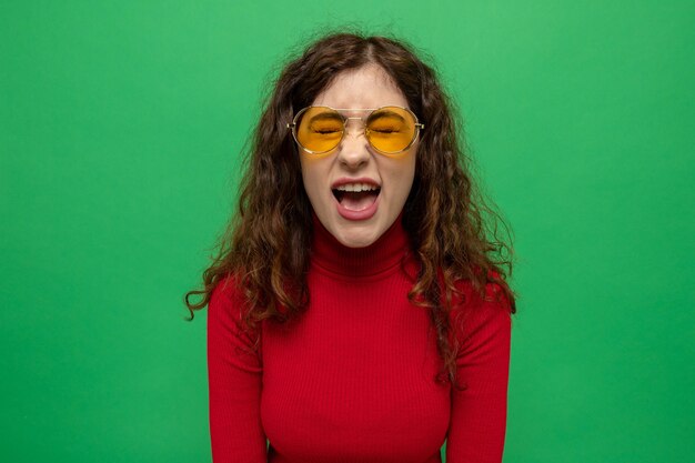 Young beautiful woman in red turtleneck wearing yellow glasses yelling being frustrated and excited