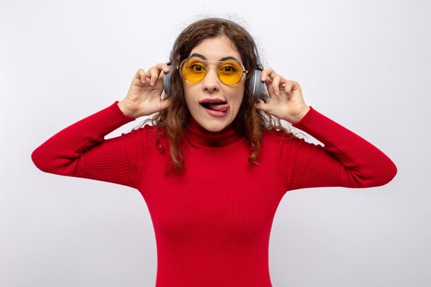 Young beautiful woman in red turtleneck with headphones wearing yellow glasses looking happy and cheerful sticking out tongue enjoying favorite music