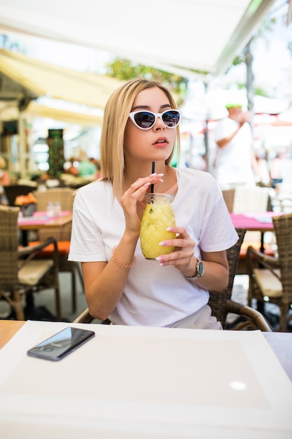 Young beautiful woman in round sunglasses with cocktail at the terrace of cafe having fun