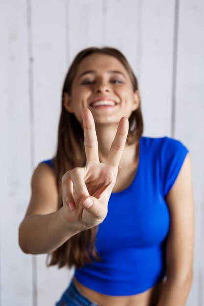 Foto gratuita giovane bella donna che mostra v gesto che sorride sopra la parete di legno bianca.