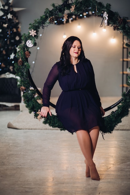 Free photo a young beautiful woman sits in a large round christmas decoration made of spruce branches