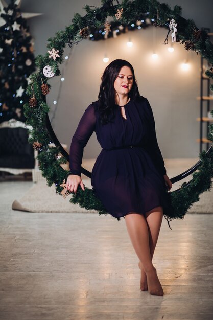 A young beautiful woman sits in a large round Christmas decoration made of spruce branches