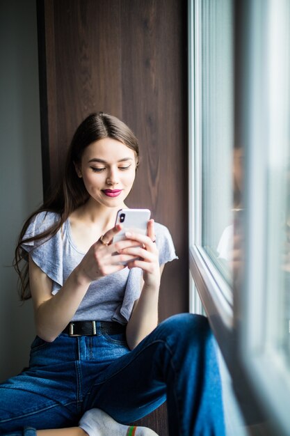 Молодая красивая женщина сидит на подоконнике на своем мобильном телефоне