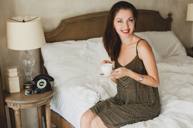 Молодая красивая женщина сидит на кровати в отеле, стильное платье, чувственное настроение, пьет кофе, держит чашку