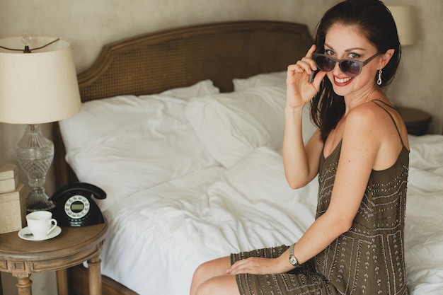 Free Photo young beautiful woman sitting on bed in hotel, stylish dress, smiling, happy, sunglasses