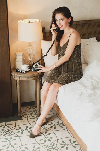 Молодая красивая женщина сидит на кровати в отеле, стильное вечернее платье, чувственное настроение, разговаривает по телефону, улыбается, кокетливо, смотрит, сексуально