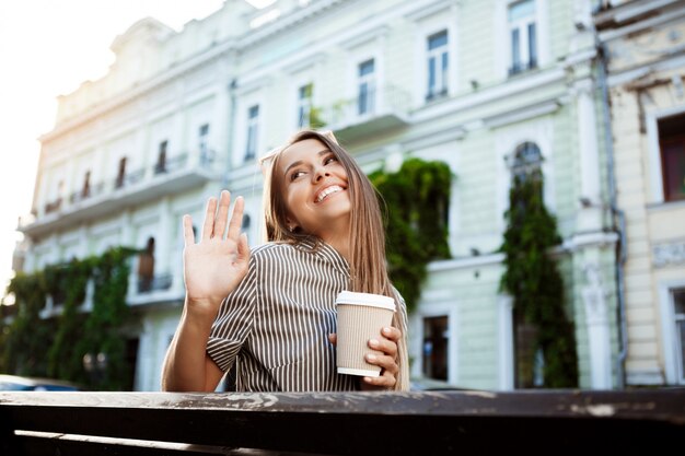 Young beautiful woman sitting on bench, holding coffee, smiling.