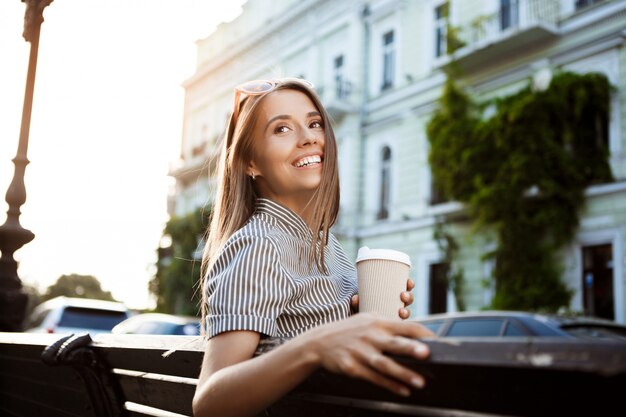 Young beautiful woman sitting on bench, holding coffee, smiling.