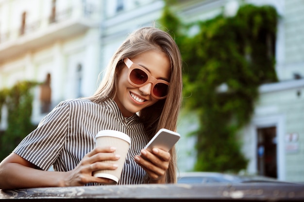 Young beautiful woman sitting on bench, looking at phone, smiling.