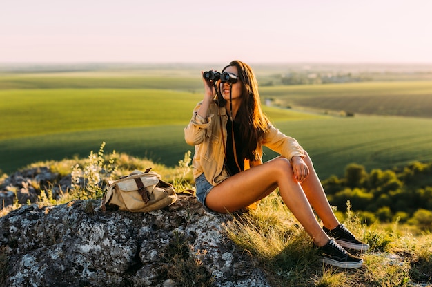 双眼鏡を見ている岩に座っている若い美しい女性