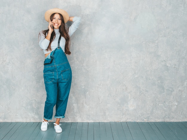 Young beautiful woman speaking on phone. Trendy girl in casual summer overalls clothes and hat. 