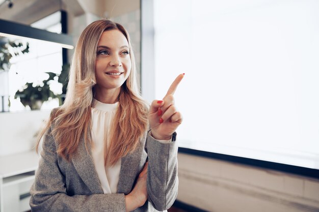 オフィスの窓の近くに立って、スペースをコピーするために指を指している若い美しい女性