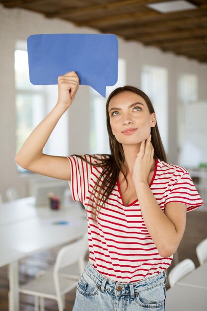 Young beautiful woman in striped Tshirt dreamily looking up holding blue paper shape of message in hand under head at work in modern empty office