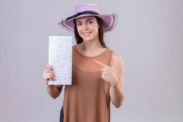 白い背景の上に立って幸せそうな顔に笑みを浮かべてそれを指している地図を保持している夏帽子の若い美しい女性