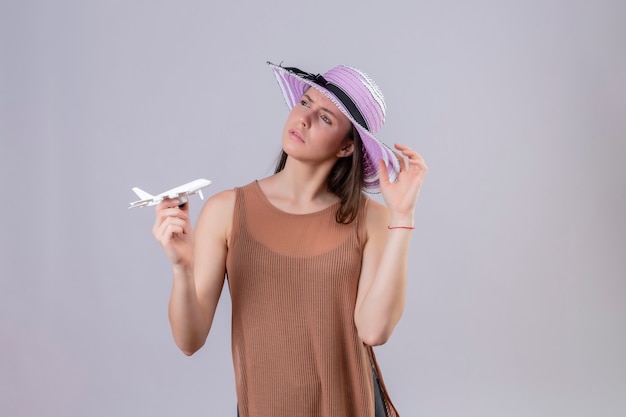 Young beautiful woman in summer hat holding toy airplane looking aside thinking with pensive expression standing over white background