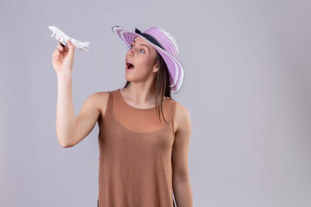 白い壁に遊び心と幸せを探しておもちゃの飛行機を保持している夏帽子の若い美しい女性