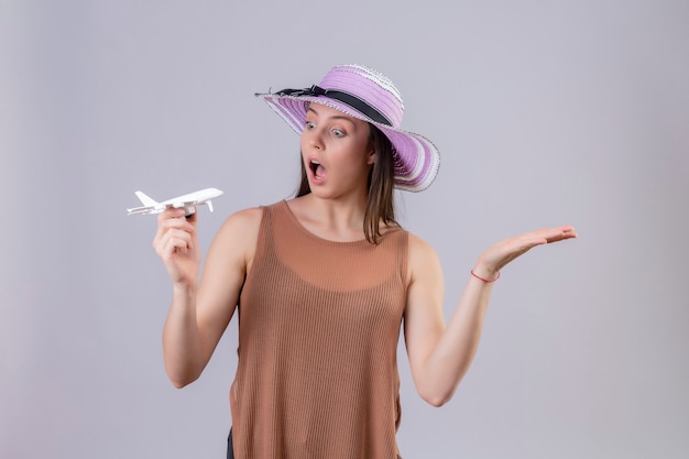 Giovane bella donna in cappello estivo che tiene aeroplano giocattolo guardando sorpreso con il braccio alzato in piedi su sfondo bianco