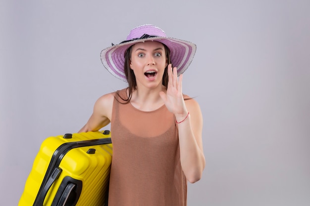 Young beautiful woman in summer hat holding yellow suitcase calling someone with hand near mouth standing over white background