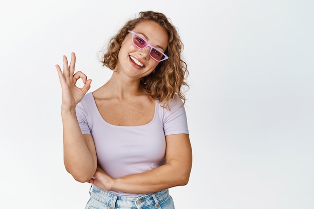 Foto gratuita la giovane bella donna in occhiali da sole, mostra il segno ok e sorride, soddisfatta di smth, raccomandando, in piedi sul bianco.