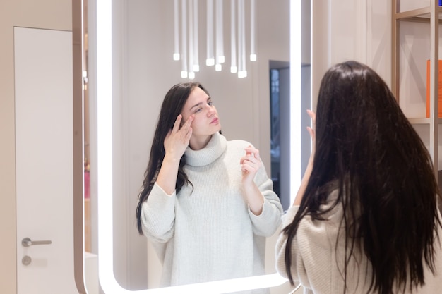 Young beautiful woman in a sweater in a beauty salon looks in the mirror, touches her face, thinks about the upcoming procedures, considers herself