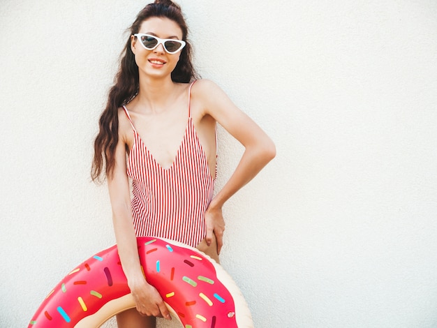 Young beautiful woman in swimwear and sunglasses posing in the street with inflatable donut