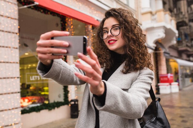 Young beautiful woman taking seflie picture using smartphone, autumn street city style, warm coat, glasses, happy, smiling, holding phone in hand, curly hair