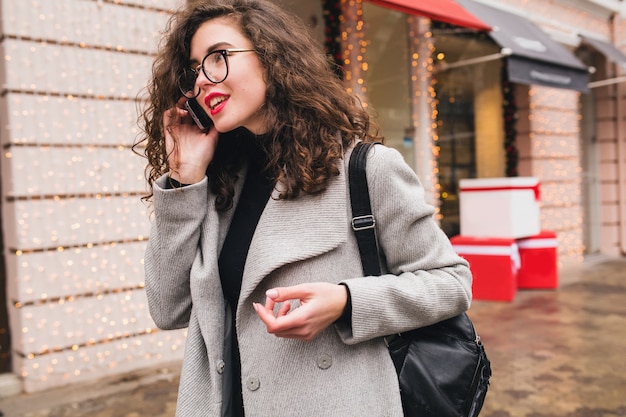Young beautiful woman talking on smartphone, autumn street city style, warm coat, glasses, happy, smiling, holding phone in hand, curly hair