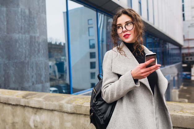 Young beautiful woman talking on smartphone, autumn street city style, warm coat, glasses, happy, smiling, holding phone in hand, curly hair