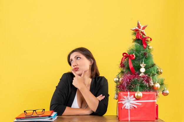 Young beautiful woman thinking about something carefully sitting at a table near decorated Christmas tree at office on yellow 