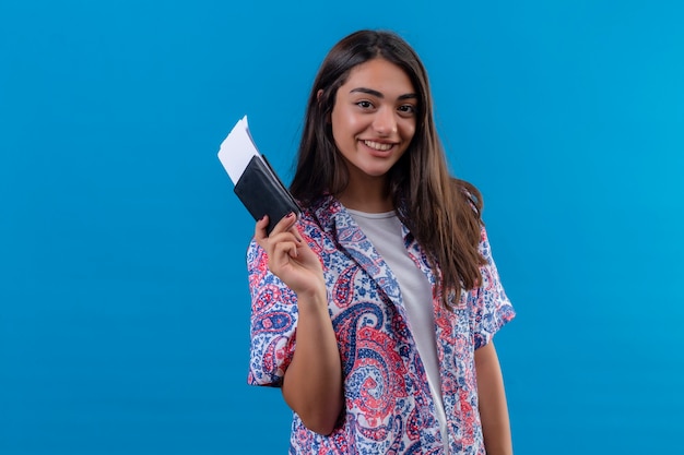 Young beautiful woman tourist holding passport with tickets looking at camera with confident smile positive and happy standing over satiated blue background
