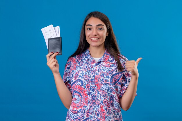 Молодая красивая женщина туристический паспорт с билетами весело улыбаясь, показывает палец вверх, готов к празднику за изолированные синей стеной