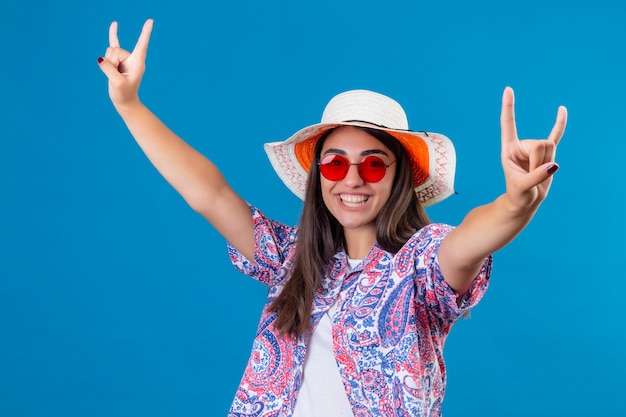 Young beautiful woman tourist wearing summer hat and red sunglasses looking joyful making rock symbols smiling cheerfully happy and positive over blue wall