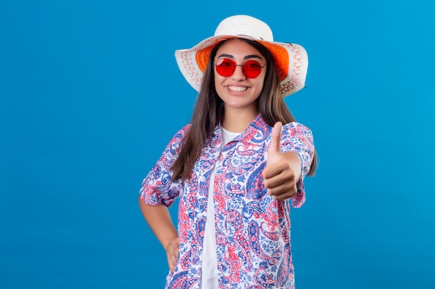 孤立した青い壁の上に親指を現して幸せそうな顔を浮かべて夏の帽子と赤いサングラスを着ている若い美しい女性観光客