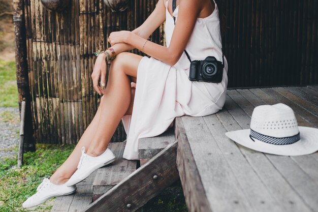 Young beautiful woman on tropical vacation in asia, summer style, white boho dress, sneakers, digital photo camera, traveler, straw hat, legs close up details