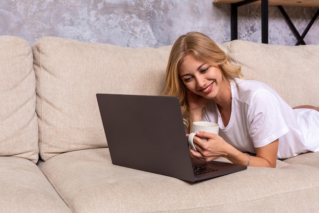 ラップトップコンピューターを使用して幸せな見て、ブラウジングまたは自宅でオンラインインターネットショッピングをしている笑顔の若い美しい女性。モダンなリビングルーム。明るく満足のいく表情
