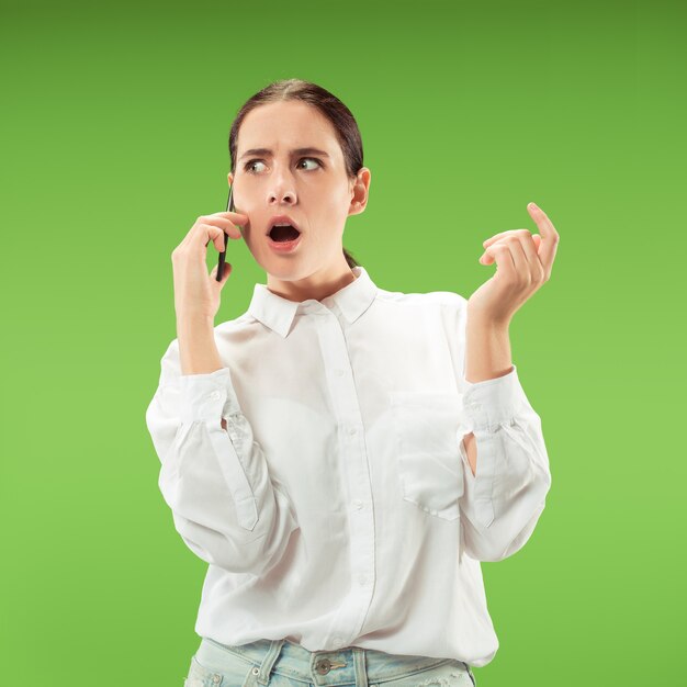 Young beautiful woman using mobile phone  on green color  wall. Human facial emotions concept. Trendy colors