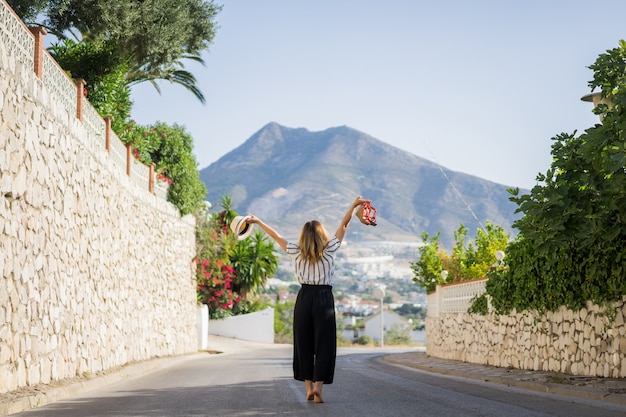 young beautiful woman on vacation jumping. In one hand sandals in the second hand hat.