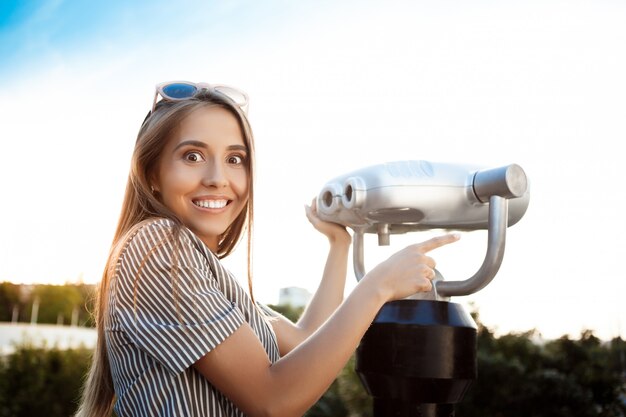 Young beautiful woman walking around city, posing near binoculars, smiling.