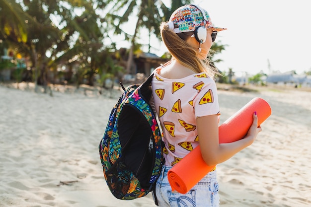 Young beautiful woman walking on beach holding yoga mat, hipster sport swag style, denim shorts, t-shirt, backpack, sunny, summer weekend