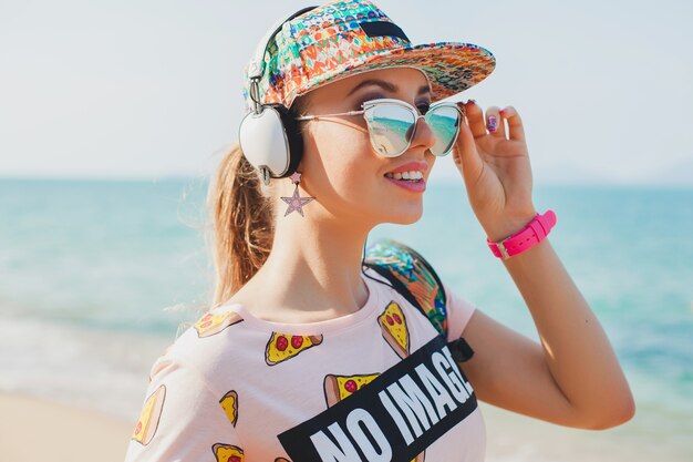 Young beautiful woman walking on beach with yoga mat, listening to music on headphones, hipster sport swag style, denim shorts, t-shirt, backpack, cap, sunglasses, sunny, summer weekend, cheerful