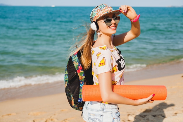 Молодая красивая женщина гуляет по пляжу с ковриком для йоги, слушает музыку в наушниках, хипстерском спортивном стиле, джинсовые шорты, футболка, рюкзак, кепка, солнцезащитные очки, солнечно, летние выходные, веселая