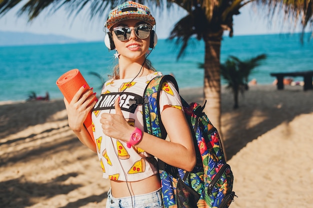 Young beautiful woman walking on beach with yoga mat, listening to music on headphones, hipster sport swag style, denim shorts, t-shirt, backpack, cap, sunglasses, sunny, summer weekend, cheerful