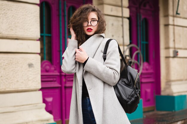 Young beautiful woman walking in the city street in grey coat, autumn fashion style, glasses, backpack,