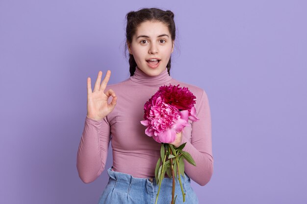 Young beautiful woman wearing casual clothing holding bouquet of roses and burgundy peony flowers, doing ok sign with fingers, excellent symbol, keeping mouth opened.