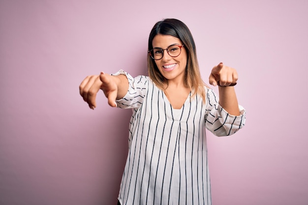 Free photo young beautiful woman wearing casual striped tshirt and glasses over pink background pointing to you and the camera with fingers smiling positive and cheerful