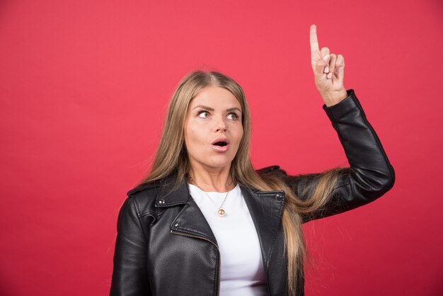 Young beautiful woman wearing leather jacket standing over red wall and pointing finger up