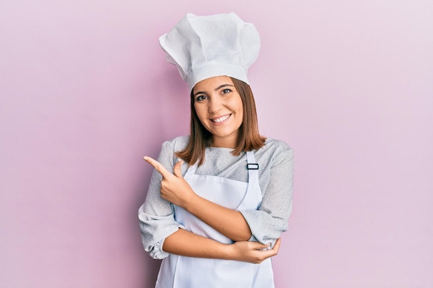 プロの料理人の制服と帽子をかぶった若い美しい女性は、顔に幸せで自然な表情を浮かべ、手と指で横を指差す顔の笑顔で陽気です