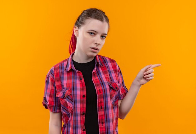  young beautiful woman wearing red shirt points finger to side on isolated yellow wall with copy space