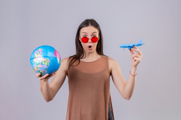 Young beautiful woman wearing red sunglasses holding globe and toy airplane amazed and surprised over white wall