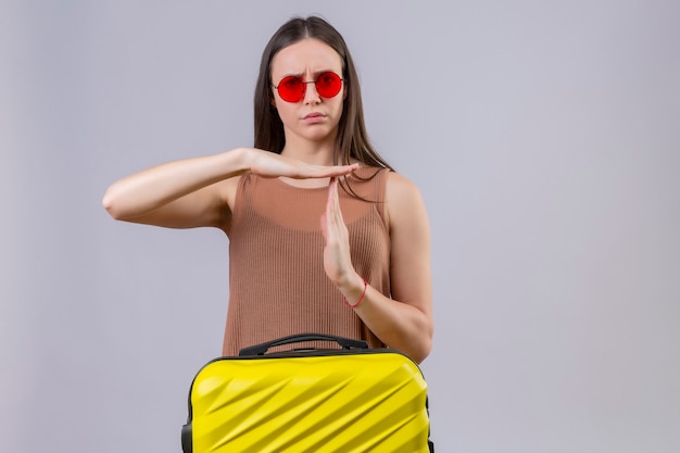 Young beautiful woman wearing red sunglasses standing with travel suitcase looking tired displeased making time out gesture with hands standing over pink background